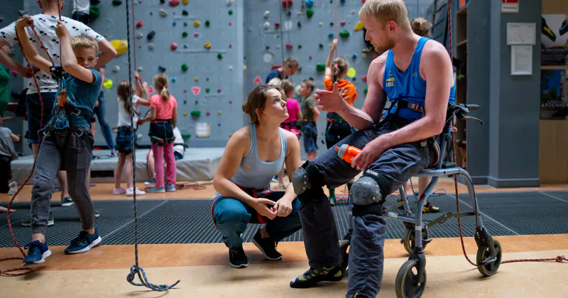 In een indoor klimhal knielt een vrouw naast een man in een rolstoel die klimuitrusting draagt, omringd door kinderen die zich ook klaarmaken om te gaan klimmen.